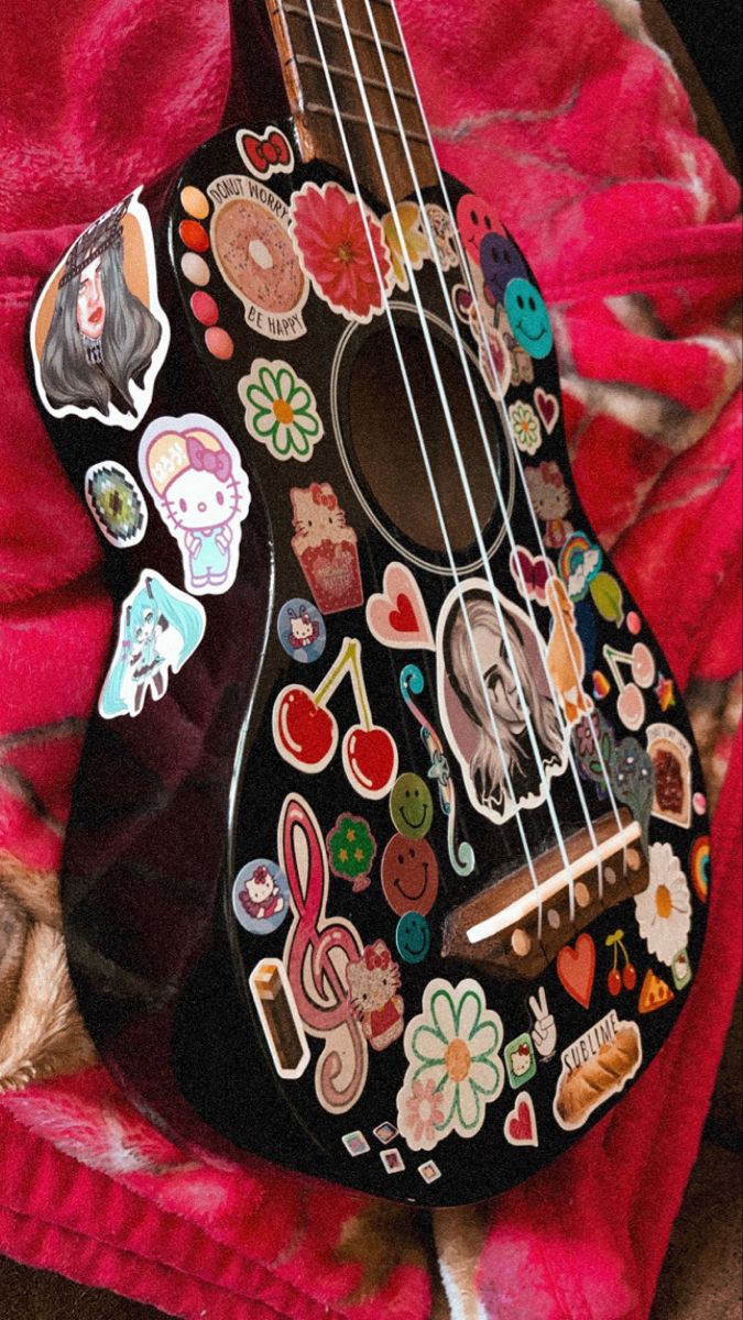 a black ukulele covered in stickers on a red blanket with a pink background