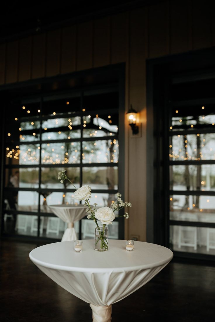 two vases with flowers on top of a white table in front of large windows