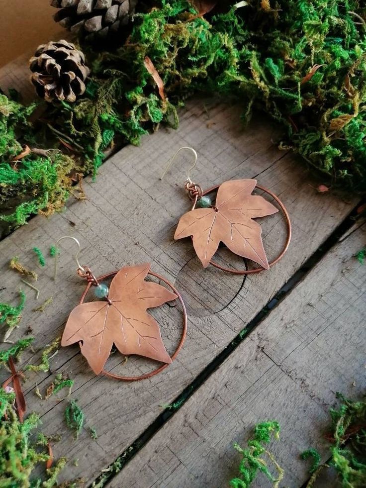 two pairs of earrings with leaves on them sitting next to some moss and pine cones