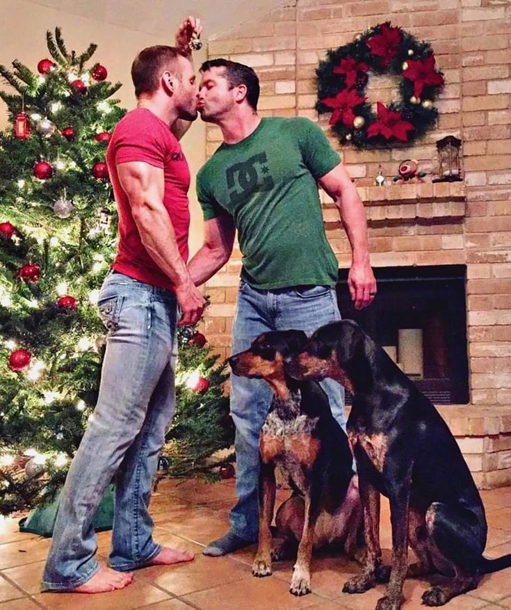 two men standing next to each other near a christmas tree with their dogs in front of them