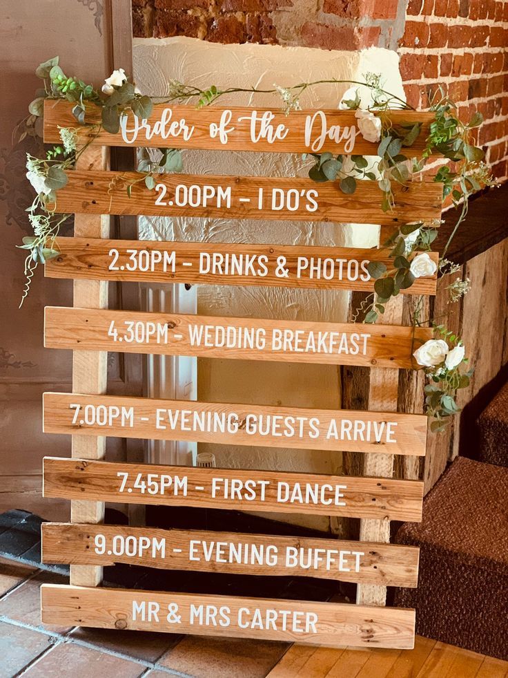 a wooden sign that says order of the day and has flowers on it in front of a brick wall