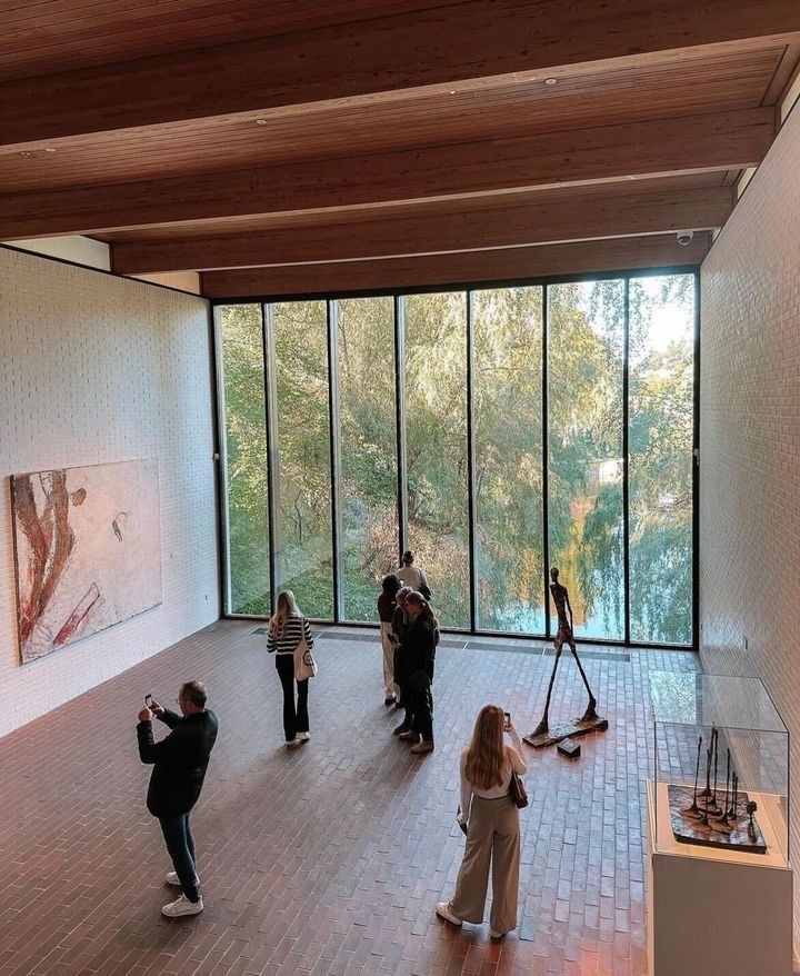 several people are standing in an open space with large windows and paintings on the walls