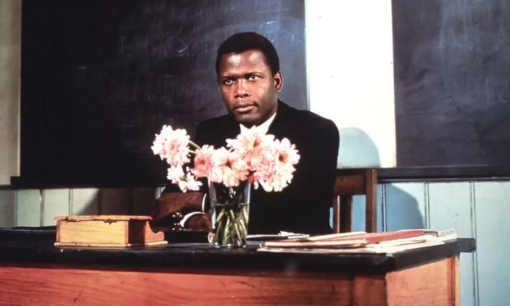 a man sitting at a desk with flowers in front of him and a blackboard behind him