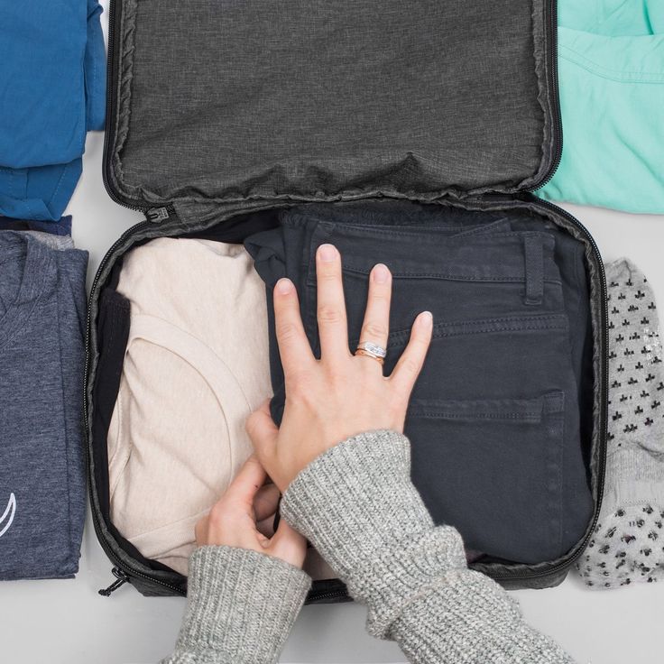 a person's hand in an open suitcase with clothes and sweaters on the floor