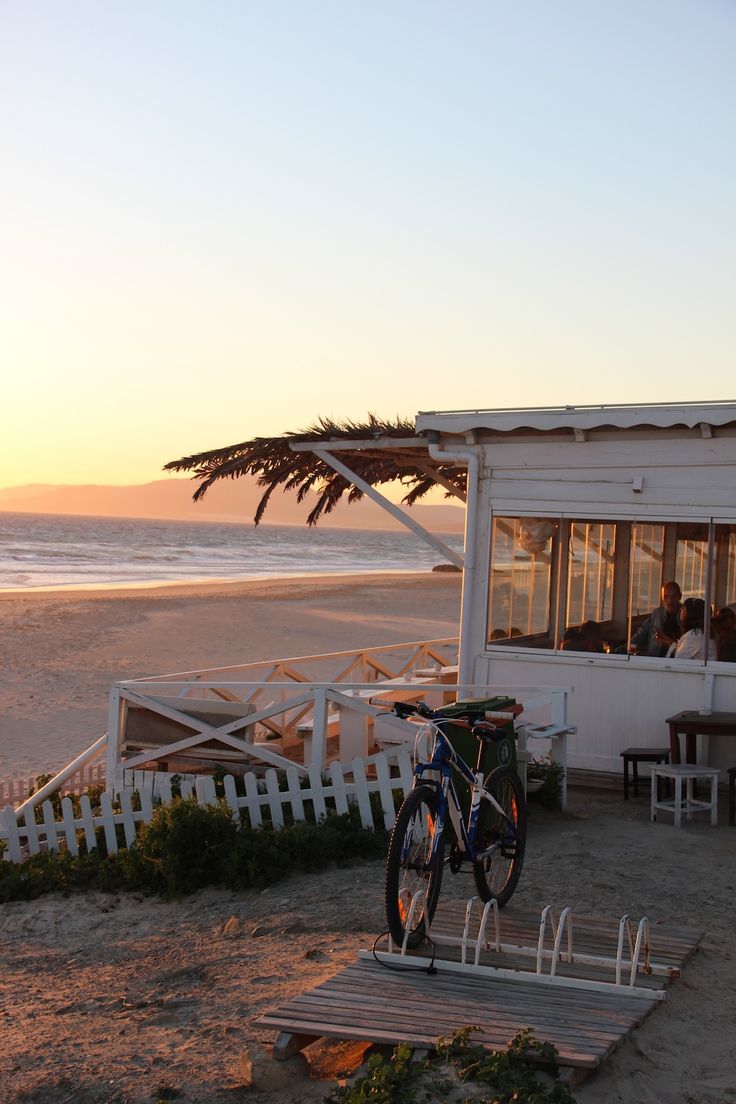 a bike parked on the beach next to a small white building with people sitting in it