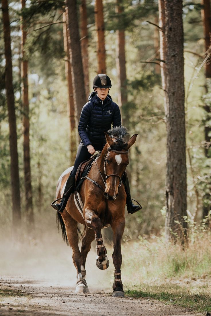 a woman riding on the back of a brown horse down a dirt road surrounded by trees