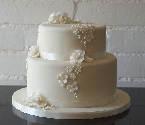 a three tiered white wedding cake with flowers on it's side, sitting on a table in front of a brick wall