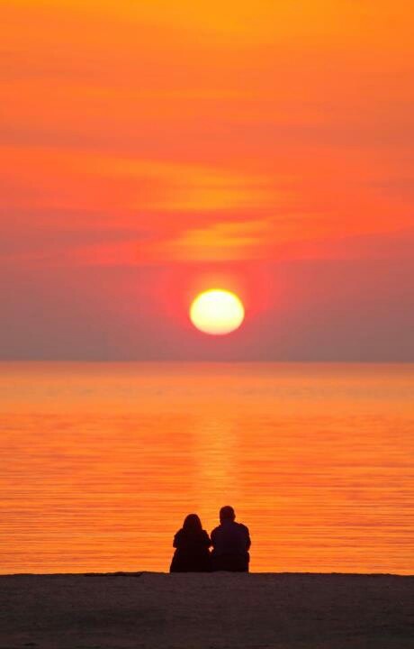 two people are sitting on the beach watching the sun go down over the water at sunset