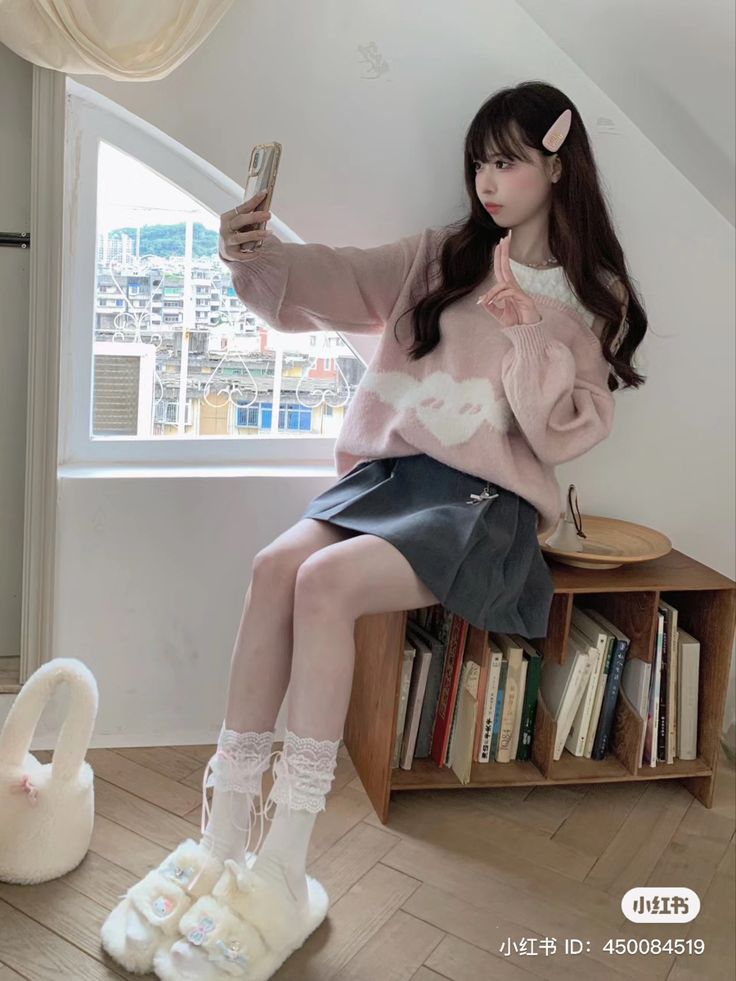 a woman sitting on top of a book shelf next to a window holding a cell phone