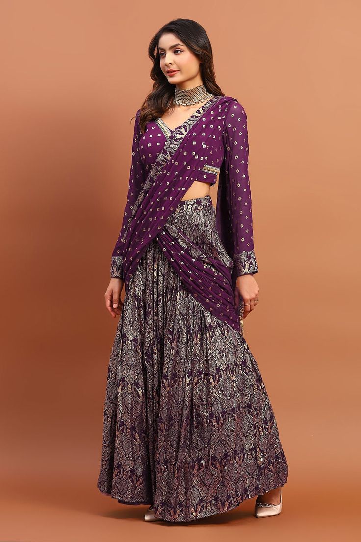 Purple georgette lehenga saree with all over vintage foil print and bandhani print attached pleated draped pallu. Paired with printed blouse with sequin and bead embroidered V neck.
Components: 2
Pattern: Printed and Hand Embroidered
Type Of Work: Bandhani, Foil Print, Bead and Sequin Work
Neckline: V Neck
Sleeve Type: Full Sleeves
Fabric: Georgette
Color: Purple
Other Details: 
Drop tasselled pallu
Pleated flare
Closure:
Saree: Side concealed zip
Blouse: Side concealed zip
Occasion: Wedding,Des Purple Pre-draped Saree For Wedding, Purple Pre-draped Saree With Zari Work For Reception, Traditional Drape Choli With Zari Work, Traditional Drape Anarkali Set With Self Design, Traditional Drape Anarkali Set For Reception, Saree-shaped Self-design Dress For Reception, Anarkali Set With Self Design For Reception, Traditional Drape Cutdana Dress For Reception, Purple Pre-draped Saree For Wedding And Eid