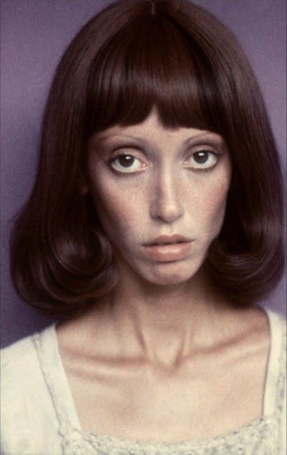 a woman with brown hair and blue eyes is posing for a photo in front of a purple background