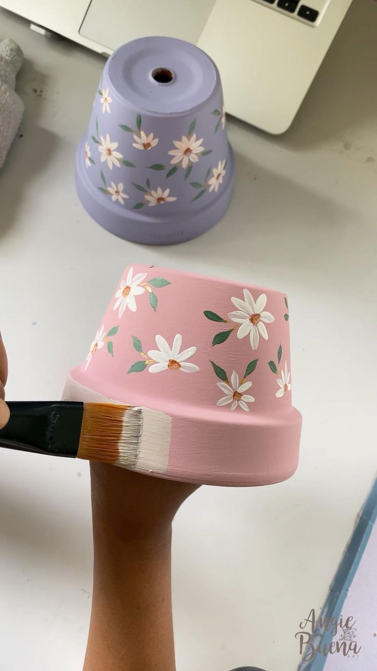 a person holding a paintbrush in front of a flowered hat on a desk