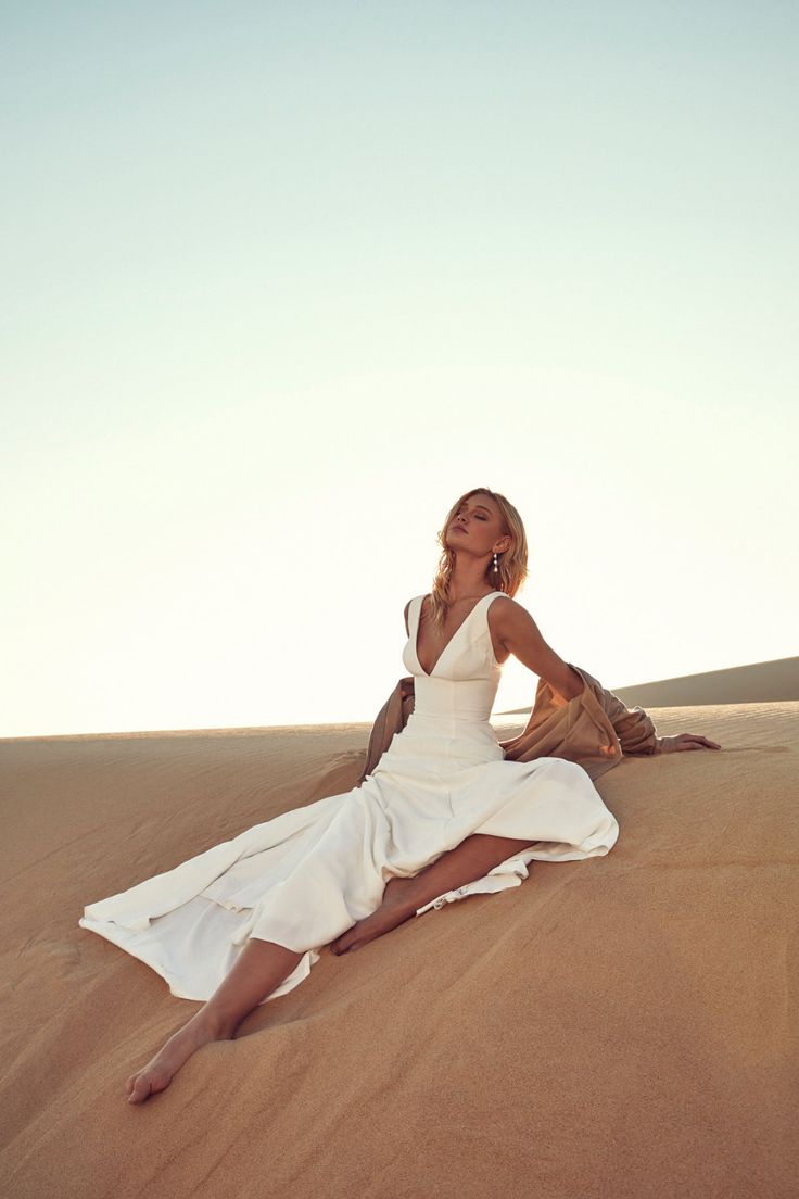 a woman in a white dress is sitting on a sand dune with her legs crossed