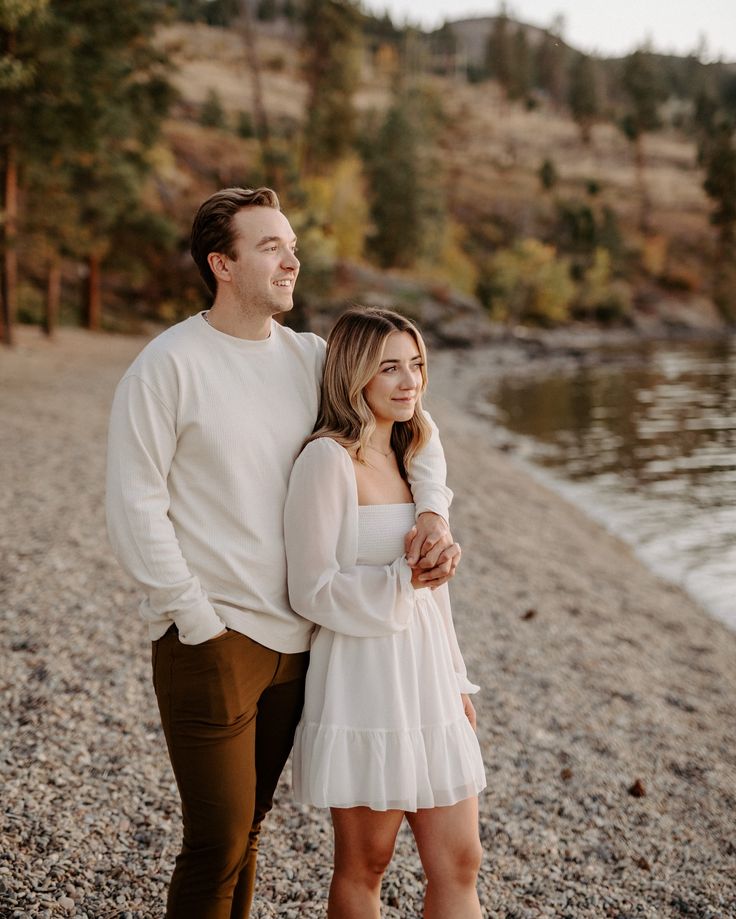 a man and woman standing next to each other on a beach near the water with trees in the background