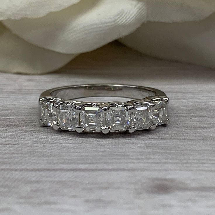 a wedding ring with five princess cut diamonds on it and a white rose in the background