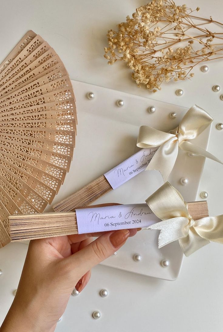 a hand holding two small wooden fan shaped tags with ribbons on them next to some flowers and pearls