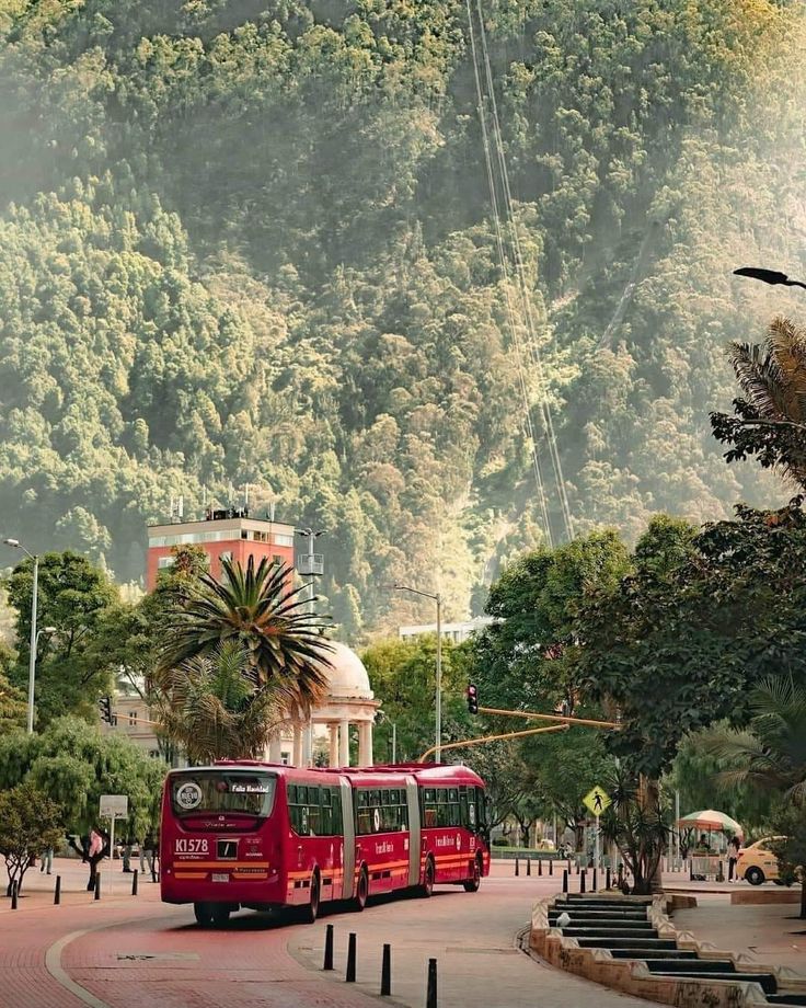 a red bus driving down a street next to a lush green forest covered mountain side