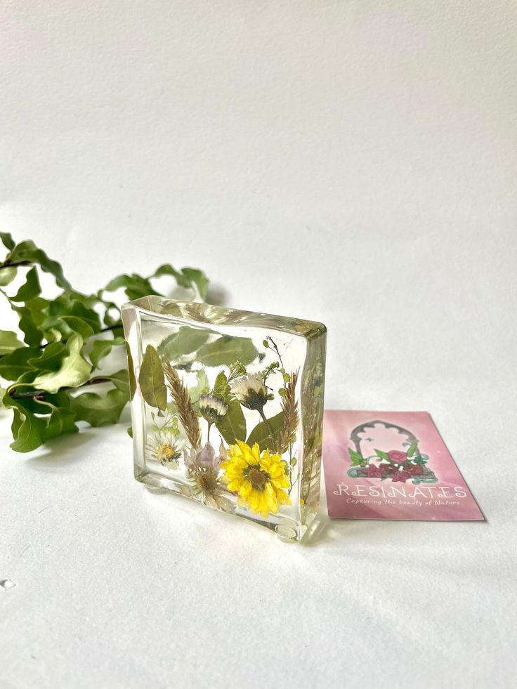 a small glass block with flowers in it and a card next to it on a white surface