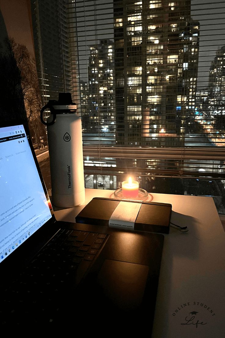 An open laptop on a desk beside a candle and planner with a nighttime cityscape in the background. Study Aesthetic Late Night, Working Late At Night Aesthetic, Night Time Routine Aesthetic Photos, Night Office Aesthetic, Writing At Night Aesthetic, Working At Night Aesthetic, College Night Aesthetic, Early Adulthood Aesthetic, Night Time Study Aesthetic