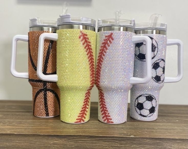 four different colored tumblers with baseball and soccer ball designs on the sides, sitting on top of a wooden table