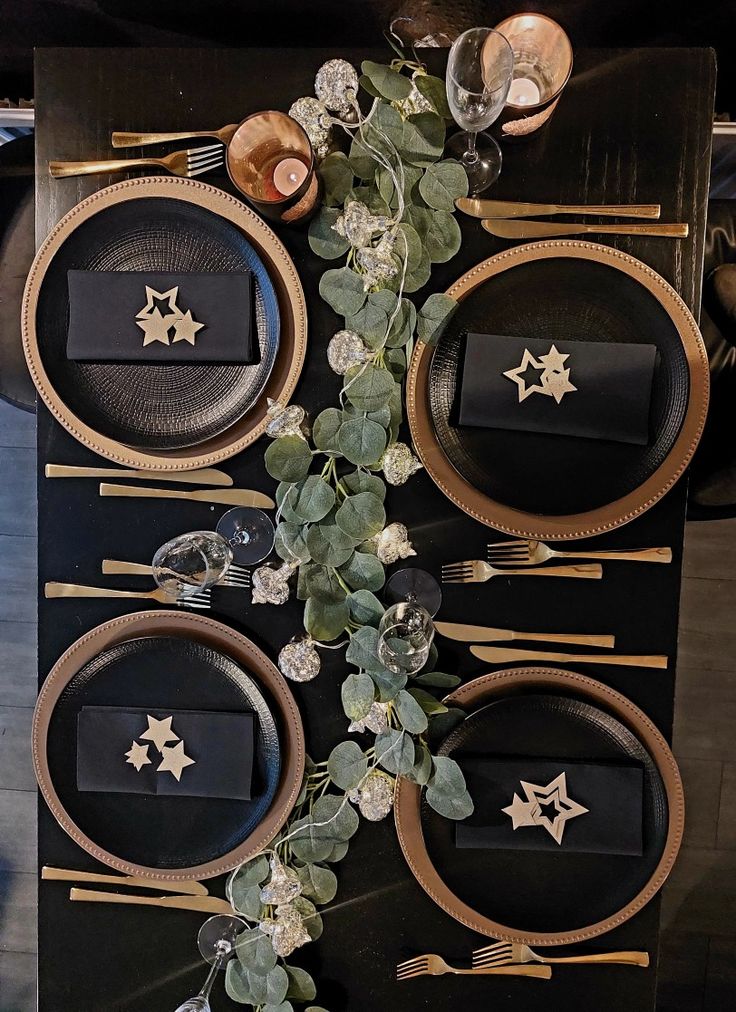 the table is set with black and gold plates, silverware, and greenery