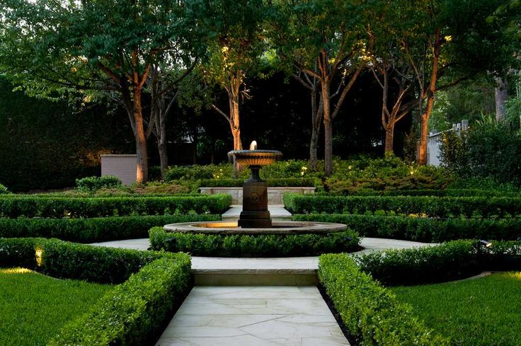 a formal garden with a fountain surrounded by hedges and trees in the background at dusk