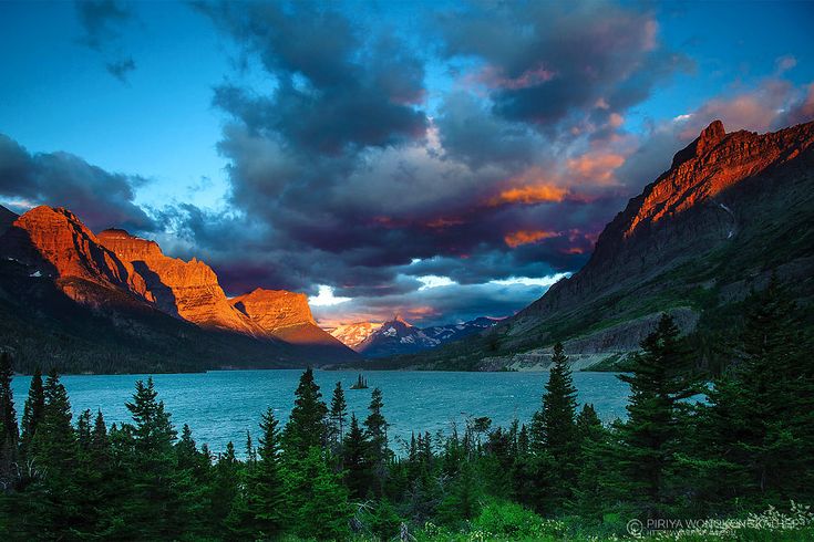 the sun is setting over some mountains and trees in front of a body of water