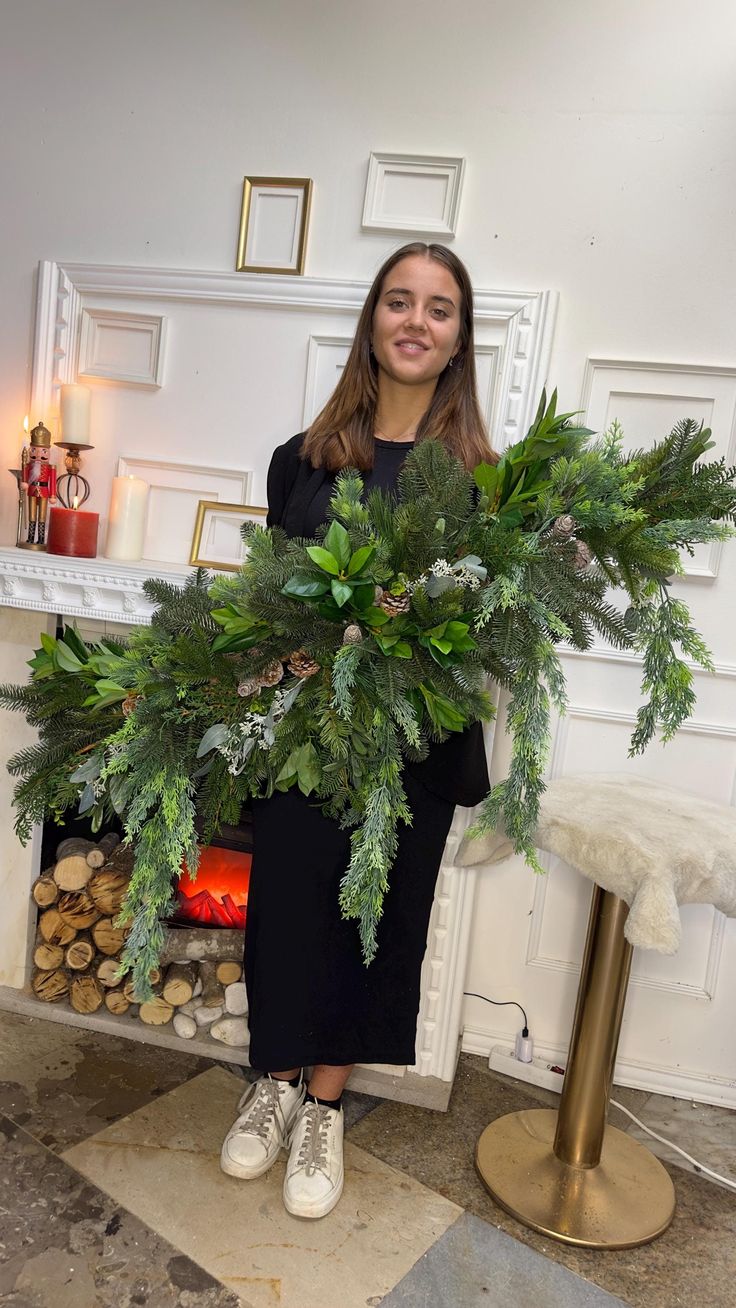 a woman standing in front of a fire place holding a large wreath with greenery on it