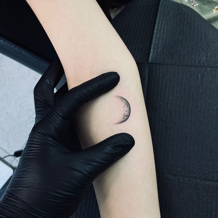 a person in black gloves holding up a small tattoo on their left arm with the moon behind it