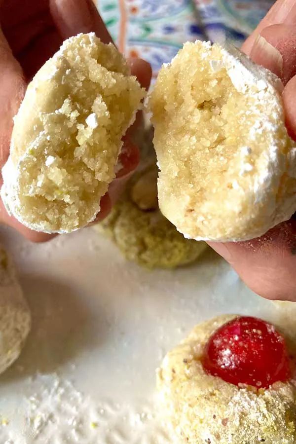 two hands are holding small pastries with powdered sugar and a cherry on top