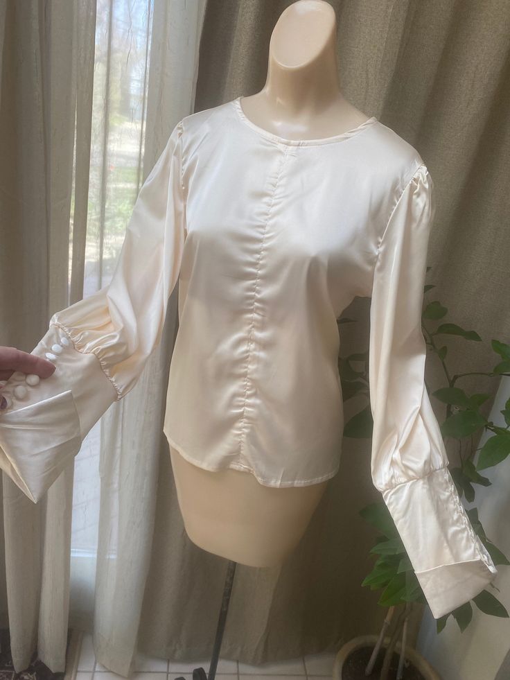 "Wonderful never worn silky feel/look blouse with the manufacturer tag still attached. Size tag says Medium but I think it would be better for a lady who wears a Small blouse from the way it fits my size Small dress form. The fabric tag in a side seam says this is 65% cotton and 35% polyester which I find strange as the fabric feels just like silk. I don't see any indication of cotton. Nevertheless the blouse is special, and anyone who sees you wearing it will assume it is fine pure silk. Can be Feminine Long Sleeve Satin Tops, Elegant Cream Long Sleeve Tops, Elegant Long Sleeve Cream Tops, Beige Long Sleeve Top With Gathered Sleeves, Elegant Beige Lantern Sleeve Top, Spring Cream Blouse With Button Cuffs, Cream Blouse With Button Cuffs For Spring, Cream Satin Top For Formal Occasions, Elegant Beige Tops With Blouson Sleeves