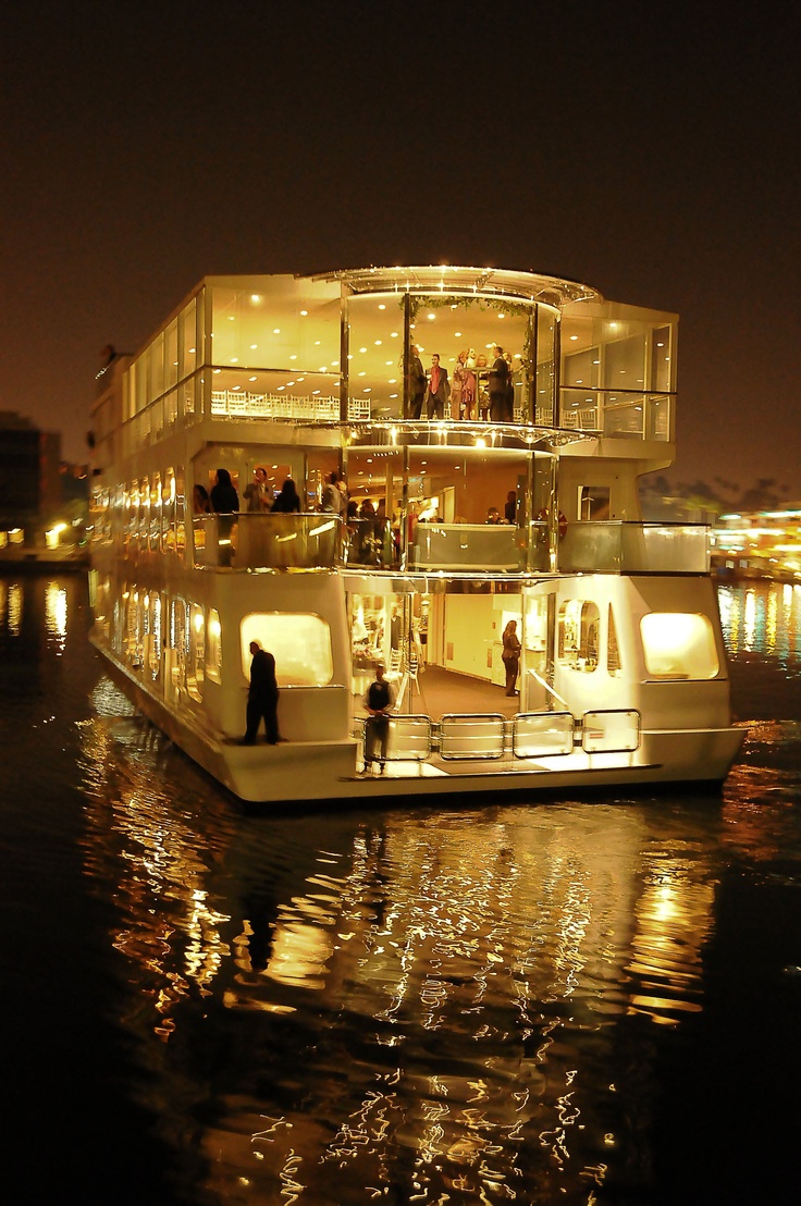 people are standing on the deck of a house boat at night in front of some buildings