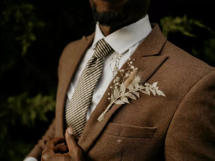 a man wearing a suit and tie with flowers on his lapel pocket, standing in front of some trees