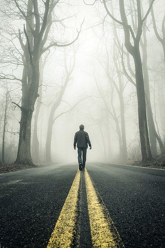 a man walking down the middle of a road with trees on both sides and fog in the background