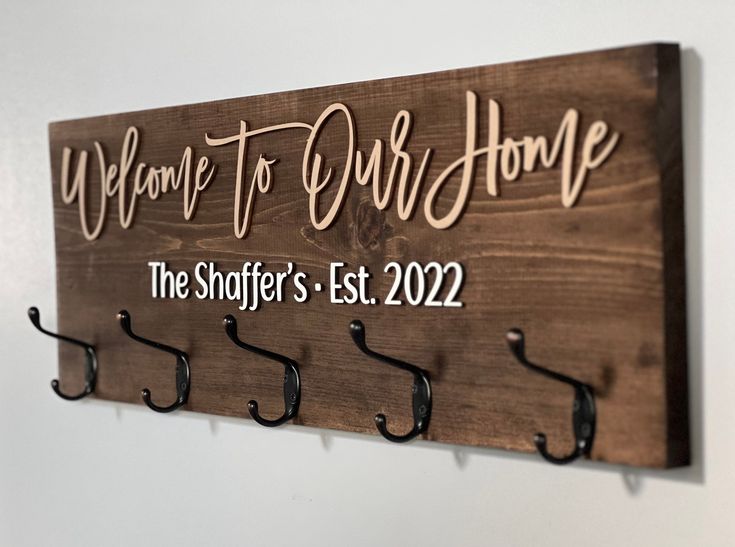 a wooden sign that says welcome to our home with hooks hanging on the wall next to it