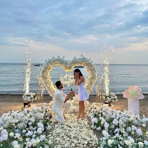 two people standing in front of a heart shaped arch with flowers on it and an umbrella