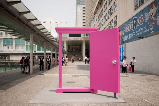 an open pink door stands in the middle of a walkway with people walking around it