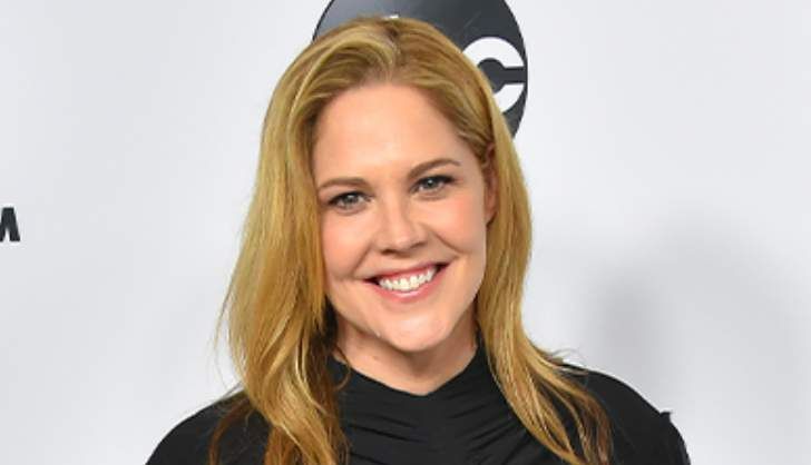 a woman with long blonde hair and black shirt smiles at the camera while standing in front of a white background