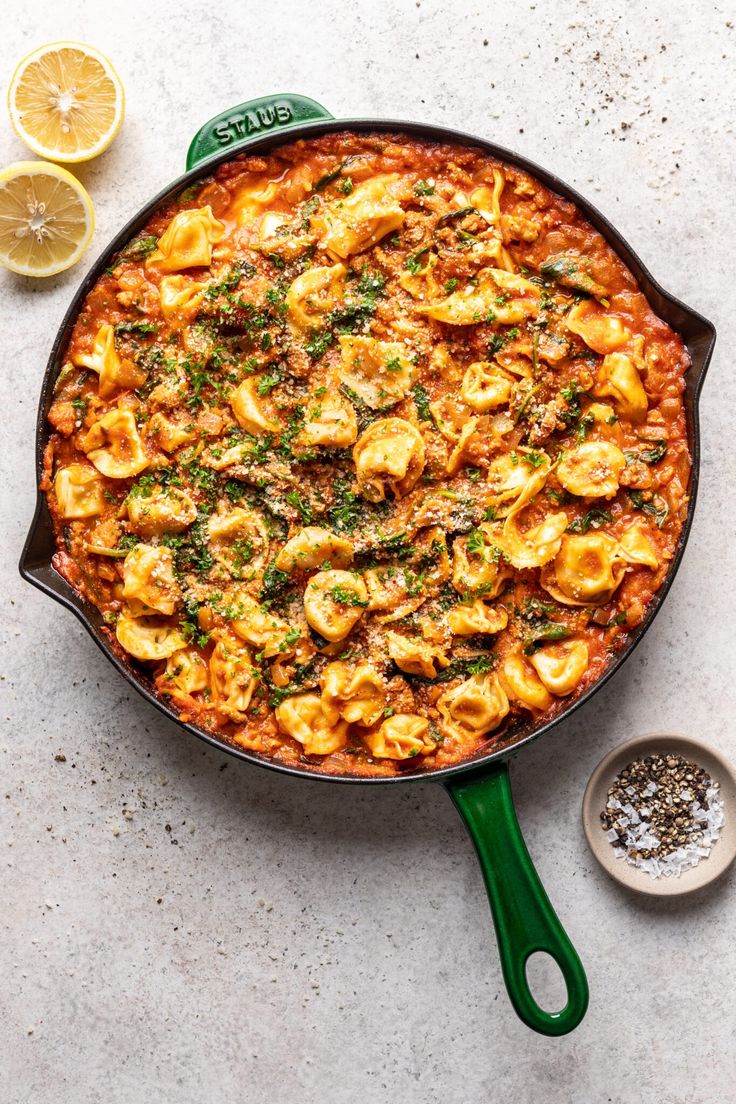 a green skillet filled with pasta and sauce next to sliced lemons on a white surface