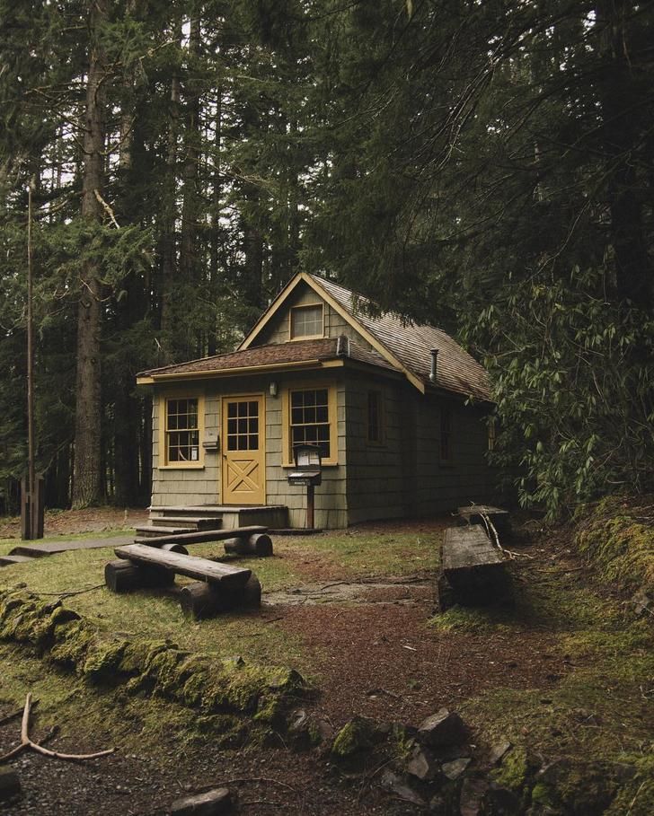 a small cabin in the middle of a forest with picnic tables and benches around it