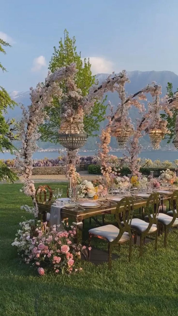 an outdoor dining table set up with flowers and greenery on the grass near water