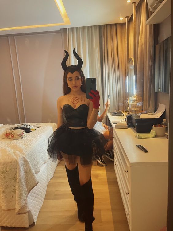 a woman in a devil costume is taking a selfie with her cell phone while standing next to a bed