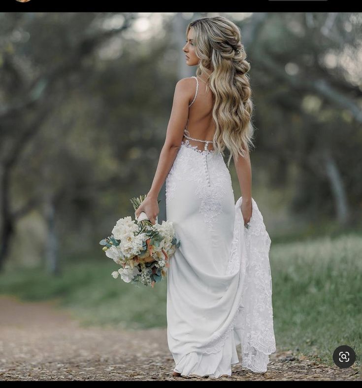 a woman in a white dress holding a bouquet and walking down a dirt road with trees behind her