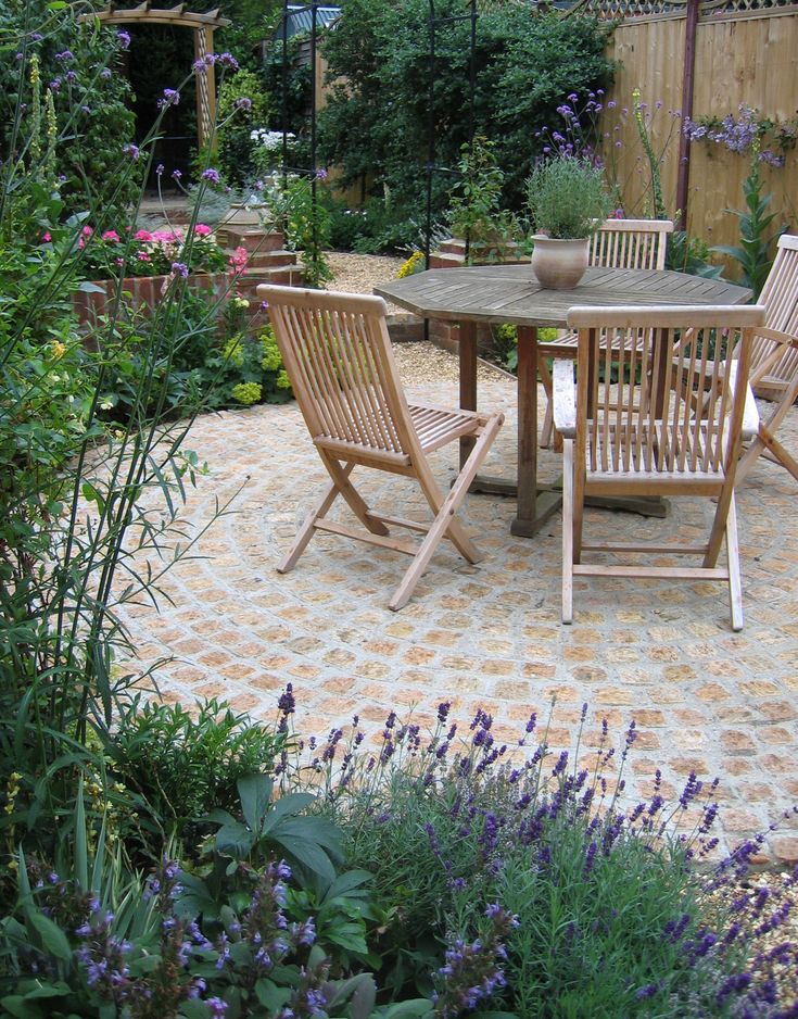 a table and chairs in a garden with purple flowers on the ground next to it