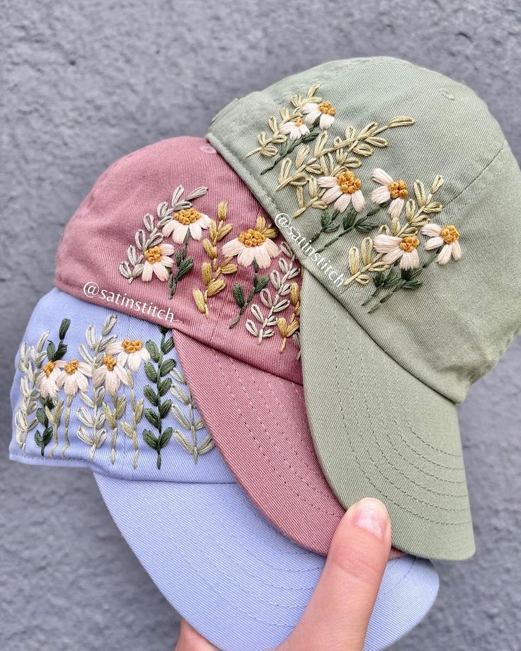 three hats with embroidered flowers on them, one being held up by a person's hand