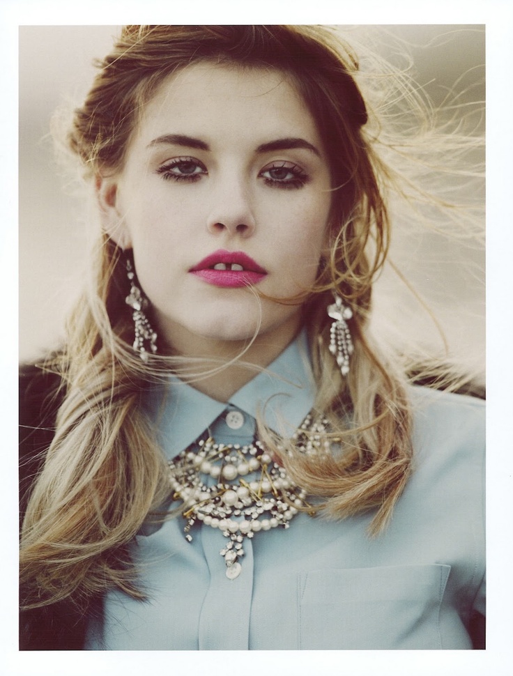 a woman with long blonde hair wearing a blue shirt and statement earrings, looking at the camera