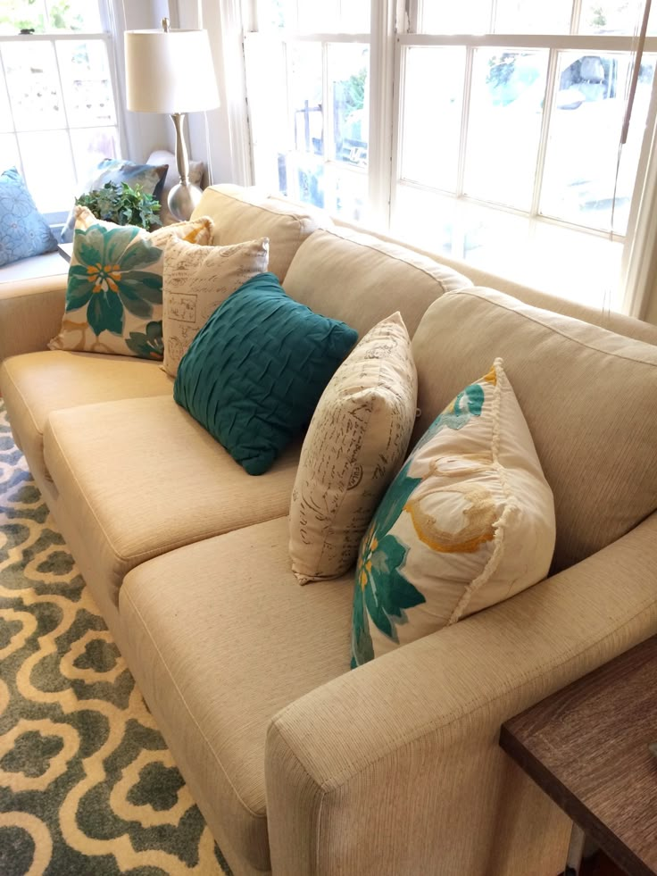 a couch with many pillows on it in front of two windows and a rug underneath the couch