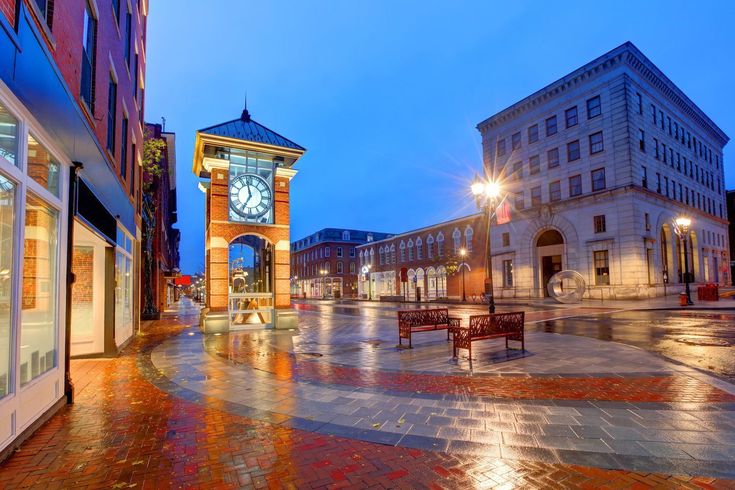 a clock tower in the middle of a town square at night with benches and tables