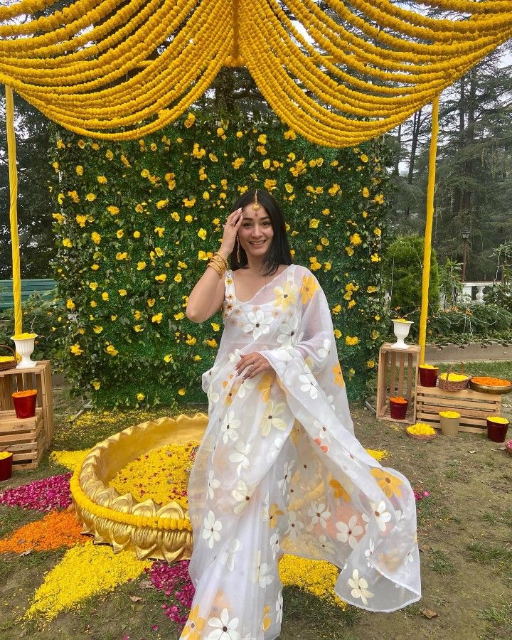 a woman in a white sari standing under a yellow flower covered structure with flowers all over it