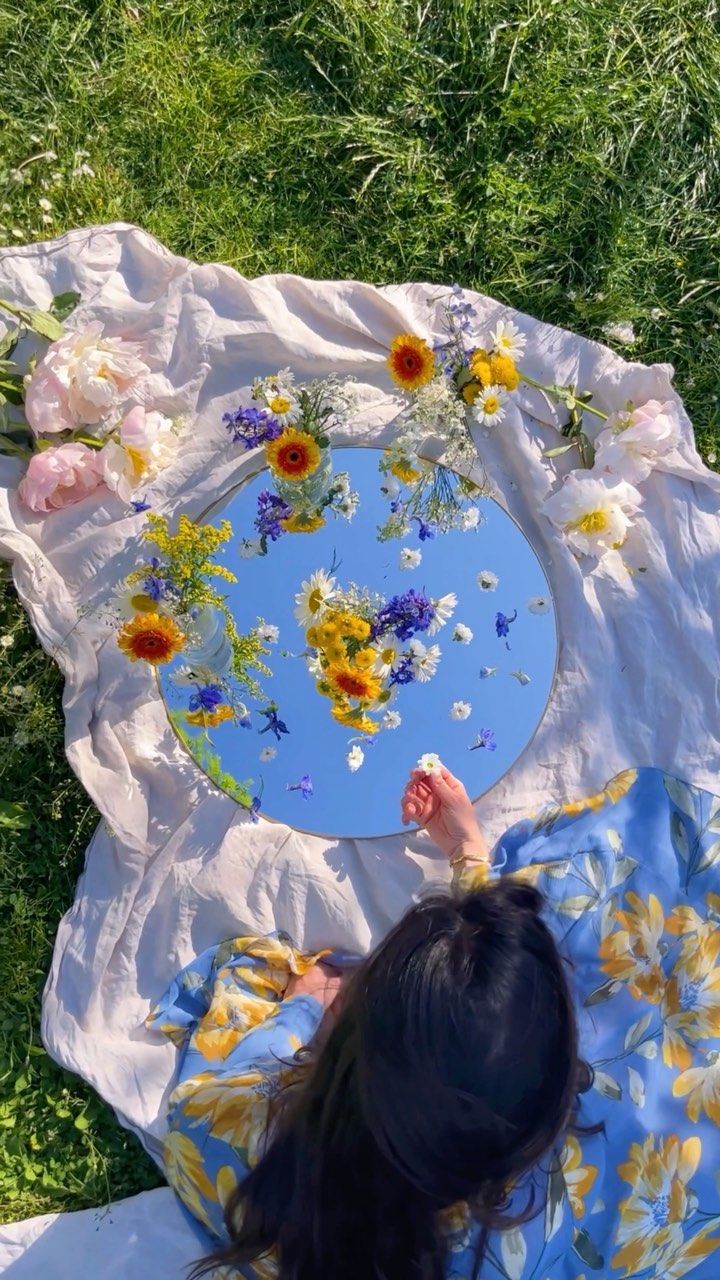 a woman laying on the ground with flowers in front of her, looking down at an upside down mirror